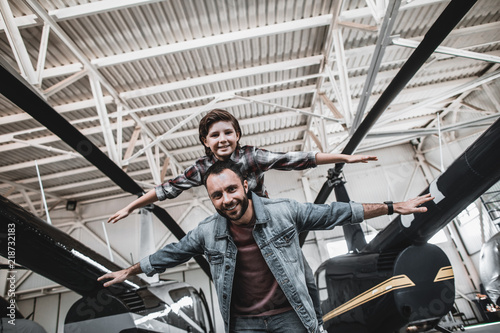 Low angle portrait of beaming bearded male playing with cheerful kid while gesticulating hands. They imagine planes while standing near rotor planes