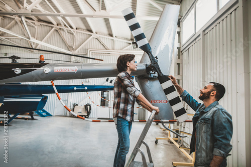 Side view smiling interested child looking at rotor of helicopter. Outgoing unshaven male twisting it while talking information about it. They locating in special garage