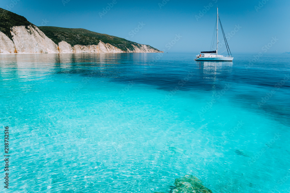 Lonely white sailing catamaran boat drift on calm sea surface. Pure shallow azure blue bay water of a beautiful beach. Scenic landscape of rocky coastline in background