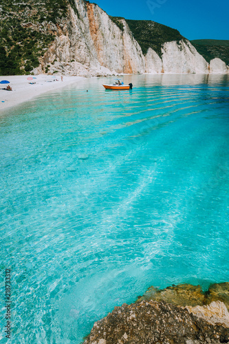 Tourist pleasure boat land to beautiful remote white pebble beach with rippled transparent clear blue sea water. Scenic landscape of rocky coastline in background