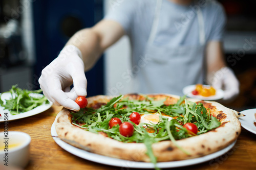 Baker in gloves putting cherry tomatoes on top of cooked pizza as decoration