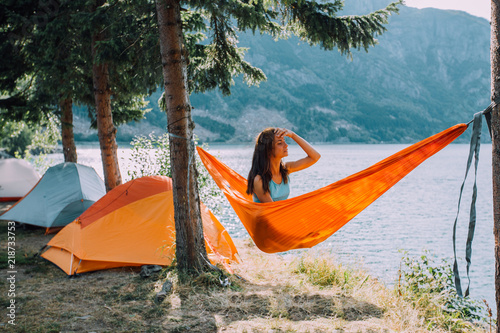 Young woman relaxing on orange hammock under trees pine enjoying the view at the lake in summer norwegian cloudy morning. Rest on camping concept.
