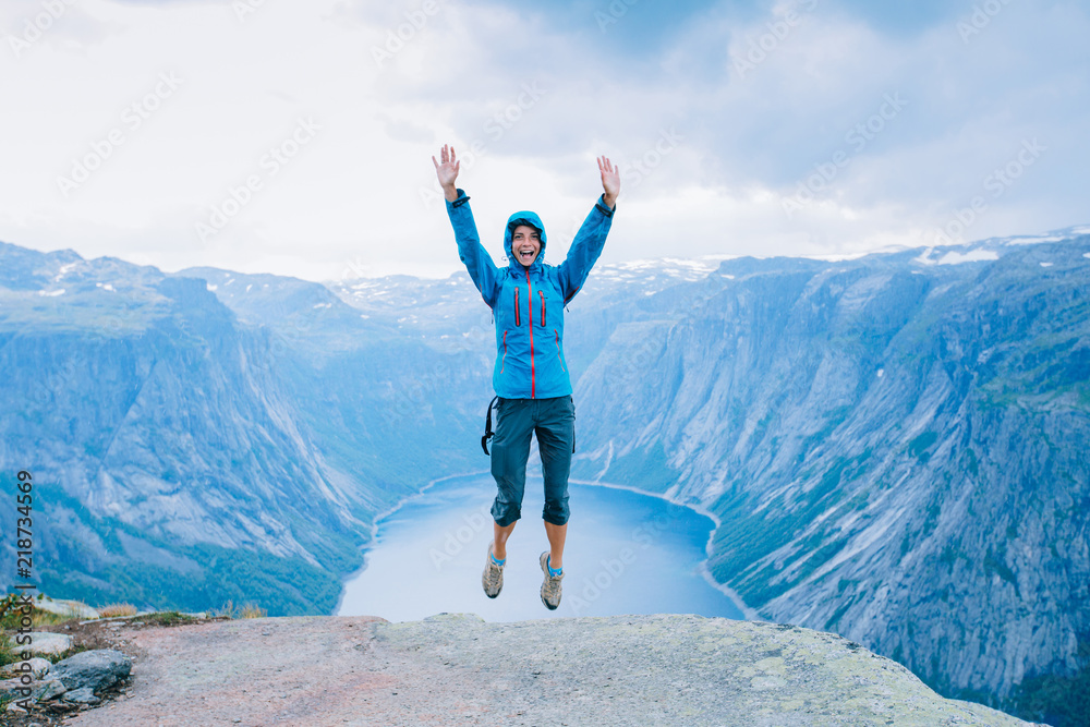 Young excited sporty woman hiker in blue jacket jumping on Norway mountain plateau. Scenic fjord on background, Norway. Young excited sporty woman hiker in blue jacket jumping on Norway mountain plateau. Scenic fjord on background, Norway.