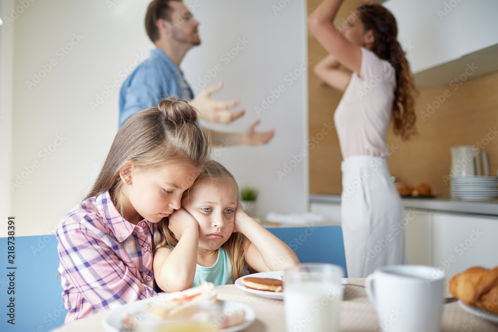 Two sad girls sitting by table in the kitchen while their parents having argument on background Two sad girls sitting by table in the kitchen while their parents having argument on background