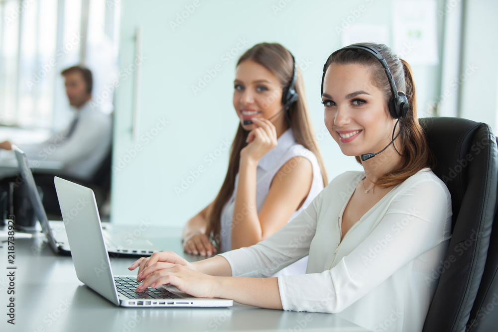 Beautiful business people in headsets are using computers and smiling while working in office. Girl is looking at camera. Beautiful business people in headsets are using computers and smiling while working in office. Girl is looking at camera.