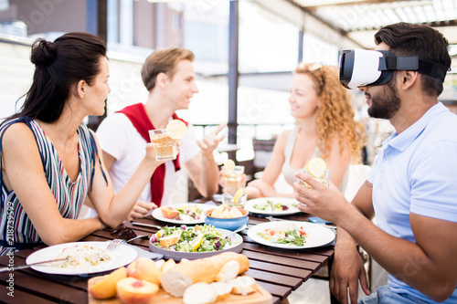 Young man in vr headset sitting by served table among his talking friends  watching video and having drink