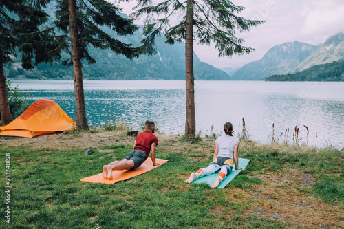Back view of sporty couple near orange tent warm up against the background of lake and mountains. The man and woman workout yoga with mats at camping in summer norwegian cloudy morning.