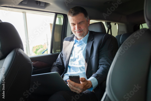Handsome businessman talking with phone sitting with laptop on the backseat of the car. © opolja