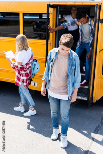 sad teenager schoolboy standing in front of school bus with classmates