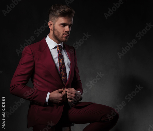 seated man buttons his red suit while looking to side