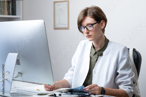 Female practitioner working at modern medical doctor office. Young medical doctor taking notes at workplace in front of a desktop computer
