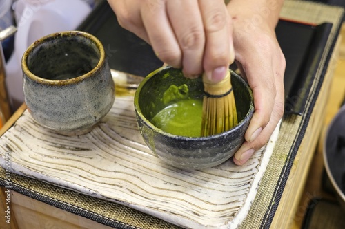 A man trying to mix a matcha green tea powder with hot water in a black bowl with a bamboo matcha whisk, demonstration of Japan green tea making art, Tools used for Japanese tea ceremony (chado).