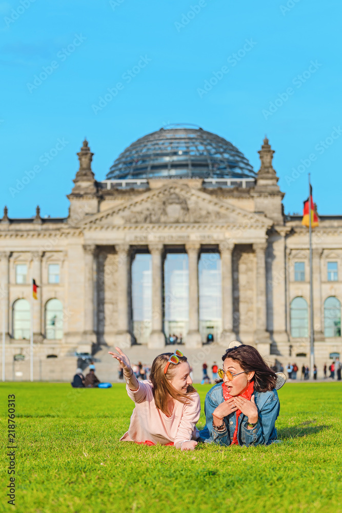 Obraz premium Dwie młode szczęśliwe dziewczyny w okularach przeciwsłonecznych leżące na trawie i bawią się przed budynkiem Bundestagu w Berlinie. Koncepcja studiów za granicą i podróży do Niemiec