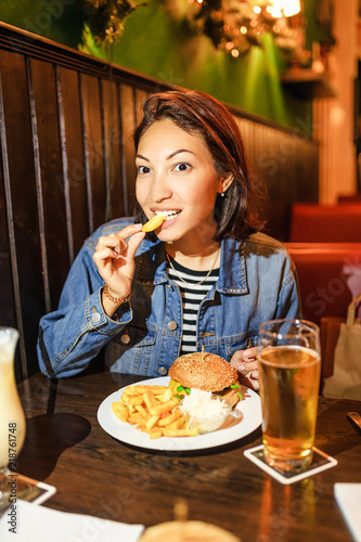 Woman eating french fries with burger and beer in pub or bar