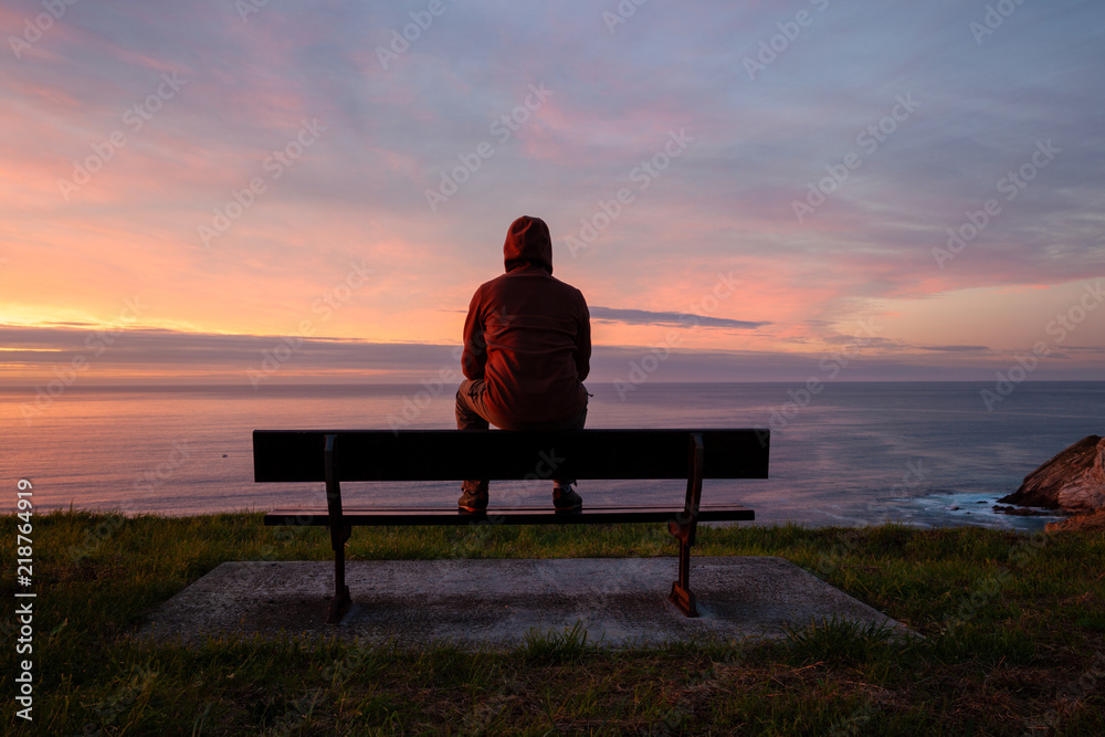 Lonely man sits alone on the rocky coast and enjoying sunset. View over rocky cliff to ocean horizon - obrazy, fototapety, plakaty 
