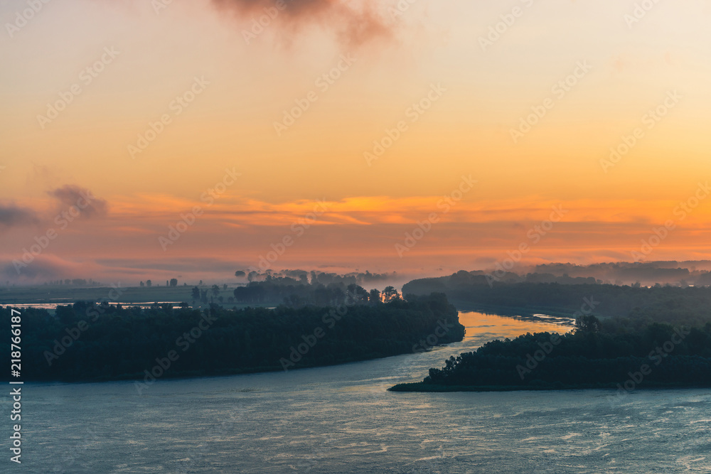 River flows along shore with forest under mist. Channel of river flows around island. Orange glow in dawn sky reflected on water. Colorful morning mystical atmospheric landscape of majestic nature.