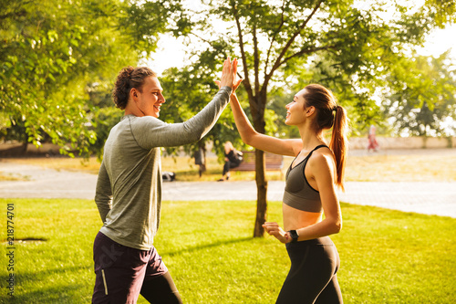 Photo of happy young man and woman 20s in tracksuits, doing workout together in green park during sunny summer day