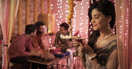 An attractive woman in a sari lighting a hanging lamp while her friends chatting in the background. A charming young lady in traditional cloths lights lamp in a decorated interior house setup photo