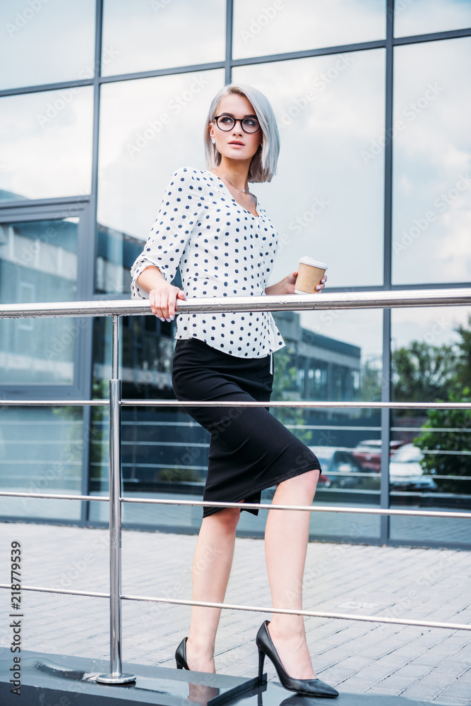 stylish businesswoman in eyeglasses with coffee to go in hand looking away on street