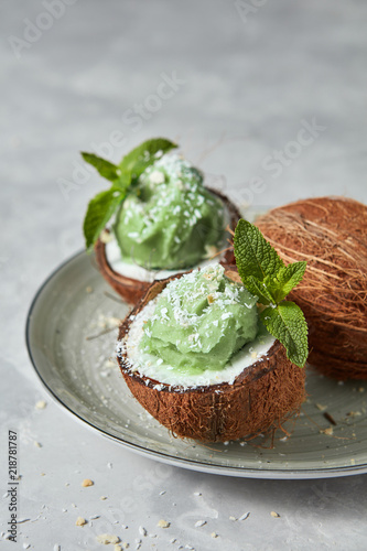 Fresh green mint ice cream in coconut shell with whole fresh coconut on a plate on a gray concrete.
