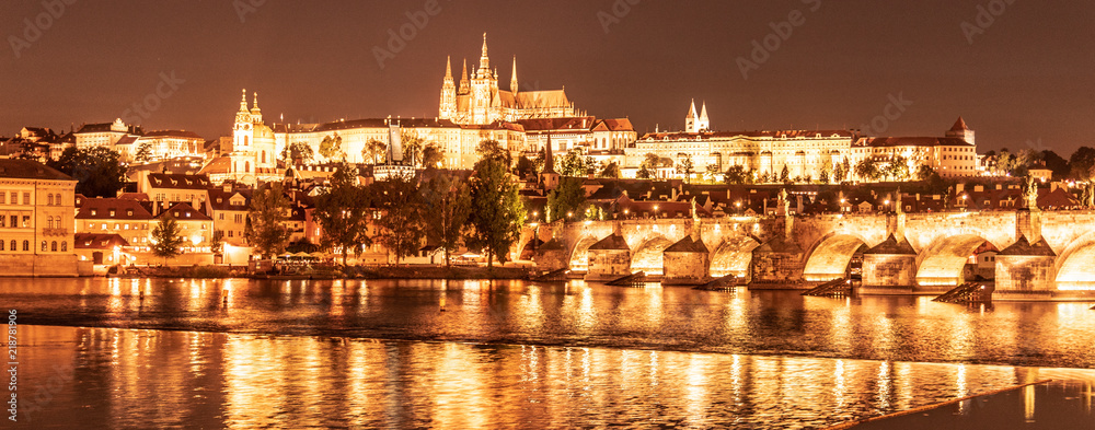 Golden Prague by night. Prague Castle and Charles Bridge reflected in Vltava River. View from Smetana Embankment. Praha, Czech Republic.