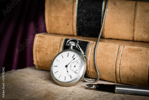 Antique vintage silver patina pocket watch with old brown leather bound books and steel fountain pen sitting on burled wood. Shallow depth of field and vignetting.