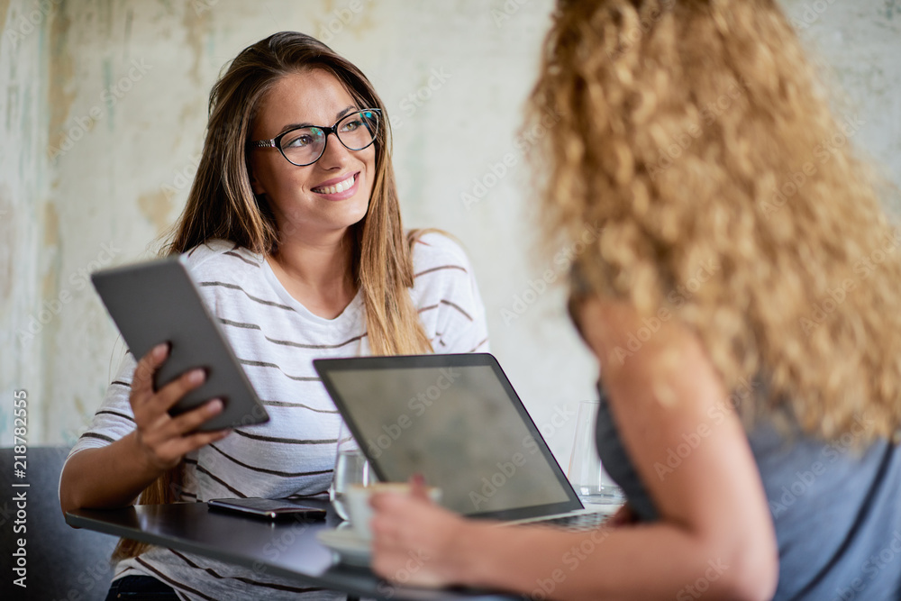 Fototapeta premium Two women using technologies and drinking coffee while sitting in cafe.