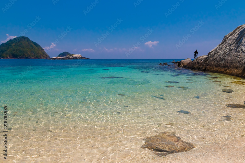 Fototapeta premium White fine sand submerged in glimmering shallow water, flanked by a big rock with a man on it & at the horizon Tukung Burung & the 2 mounds of the Susu Daras can be seen on Rawa beach, near Perhentian