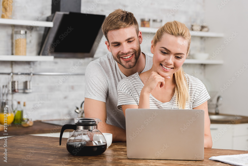 happy boyfriend hugging girlfriend in kitchen and they looking at laptop happy boyfriend hugging girlfriend in kitchen and they looking at laptop