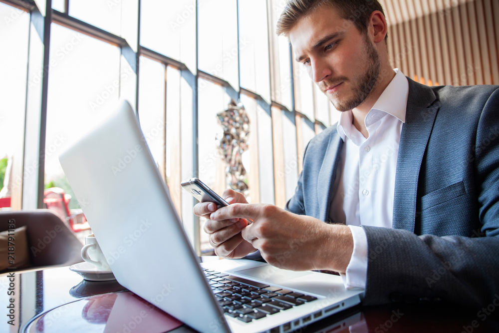 © opolja - Businessman in black suit using mobile smart phone and working on laptop computer, browsing internet and writing on paper notebook in modern office. Man working on electronics devices with copy space