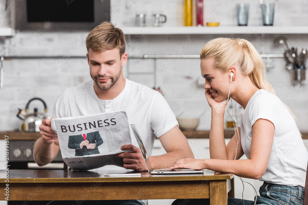 handsome young man reading business newspaper and smiling girlfriend in earphones using laptop at home handsome young man reading business newspaper and smiling girlfriend in earphones using laptop at home