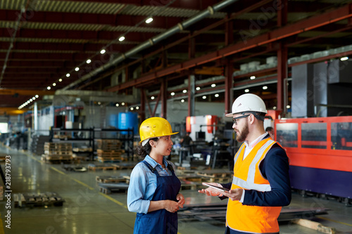 Side view portrait of production manager wearing hardhat talking to female factory worker standing in production workshop, copy space
