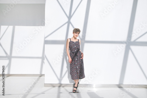 Portrait of attractive woman in dress in full height against white background with shadows. Fashionable woman in a stylish dark dress poses on the camera indoors on a white abstract background.