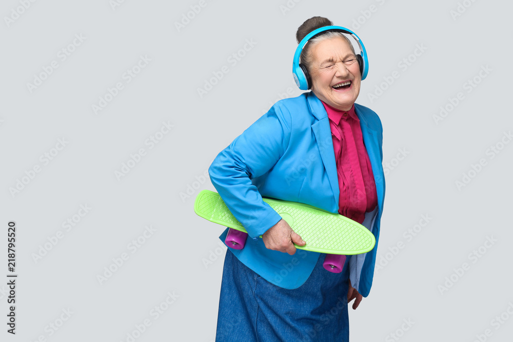 Trendy funny joyful grandmother in colorful casual style with blue headphones holding green skateboard listening music and laughing with closed eyes. indoor studio shot, isolated on gray background. - obrazy, fototapety, plakaty 