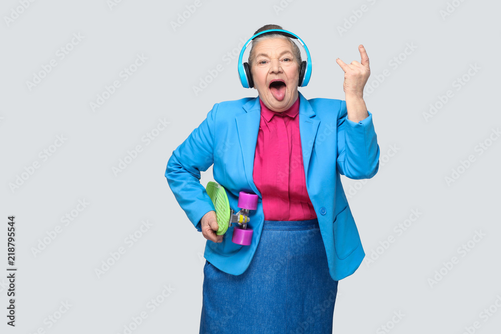 Trendy funny grandmother in casual style with blue headphones holding green skateboard listen music showing rock sign tougue out, looking at camera. Indoor, studio shot, isolated on gray background Trendy funny grandmother in casual style with blue headphones holding green skateboard listen music showing rock sign tougue out, looking at camera. Indoor, studio shot, isolated on gray background