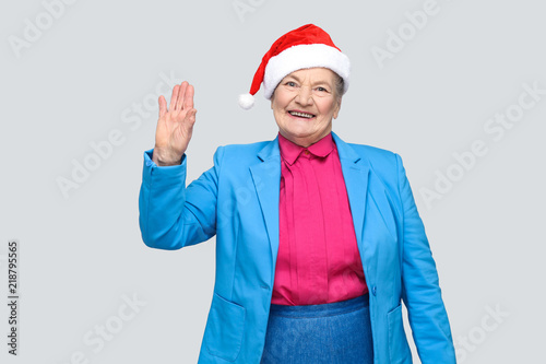 Hi, nice to see you. happy toothy smiling colorful casual style aged woman with blue suit and christmas santa red cap standing and looking at camera. indoor, studio shot, isolated on gray background