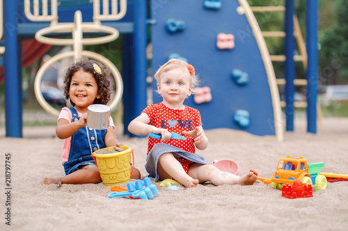 Good day. Two cute Caucasian and hispanic latin babies children sitting in sandbox playing with plastic colorful toys. Little girls friends having fun together on playground.