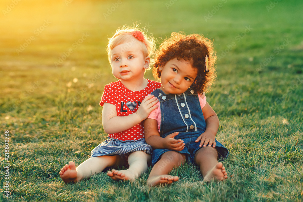 Group portrait of two cute adorable girls toddlers children sitting together. White Caucasian and latin hispanic babies hugging outside in park. Friendship and best friends forever concept. Group portrait of two cute adorable girls toddlers children sitting together. White Caucasian and latin hispanic babies hugging outside in park. Friendship and best friends forever concept.