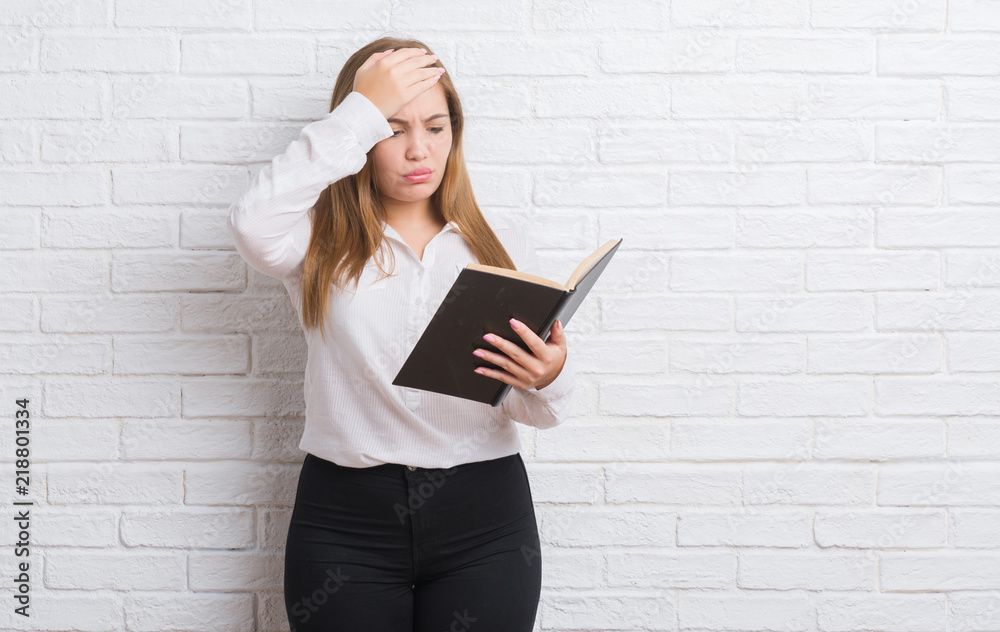 Young adult woman standing over white brick wall reading a book stressed with hand on head, shocked with shame and surprise face, angry and frustrated. Fear and upset for mistake. Young adult woman standing over white brick wall reading a book stressed with hand on head, shocked with shame and surprise face, angry and frustrated. Fear and upset for mistake.