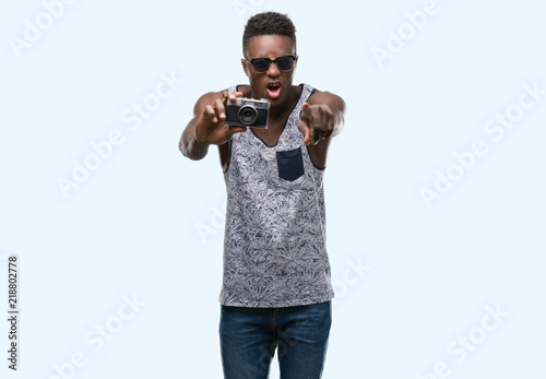 Young african american man holding vintage camera pointing with finger to the camera and to you, hand sign, positive and confident gesture from the front