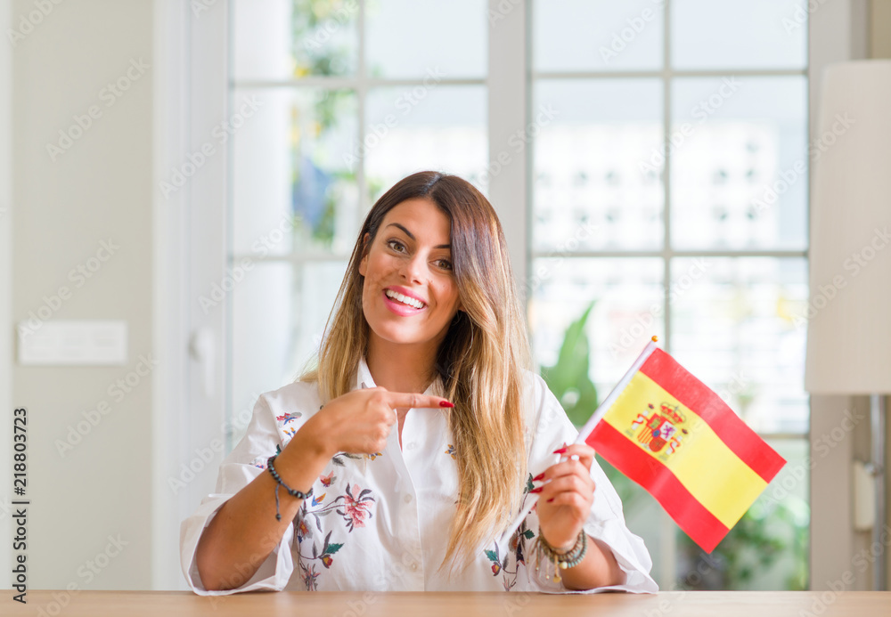 Young woman at home holding flag of Spain very happy pointing with hand and finger Young woman at home holding flag of Spain very happy pointing with hand and finger