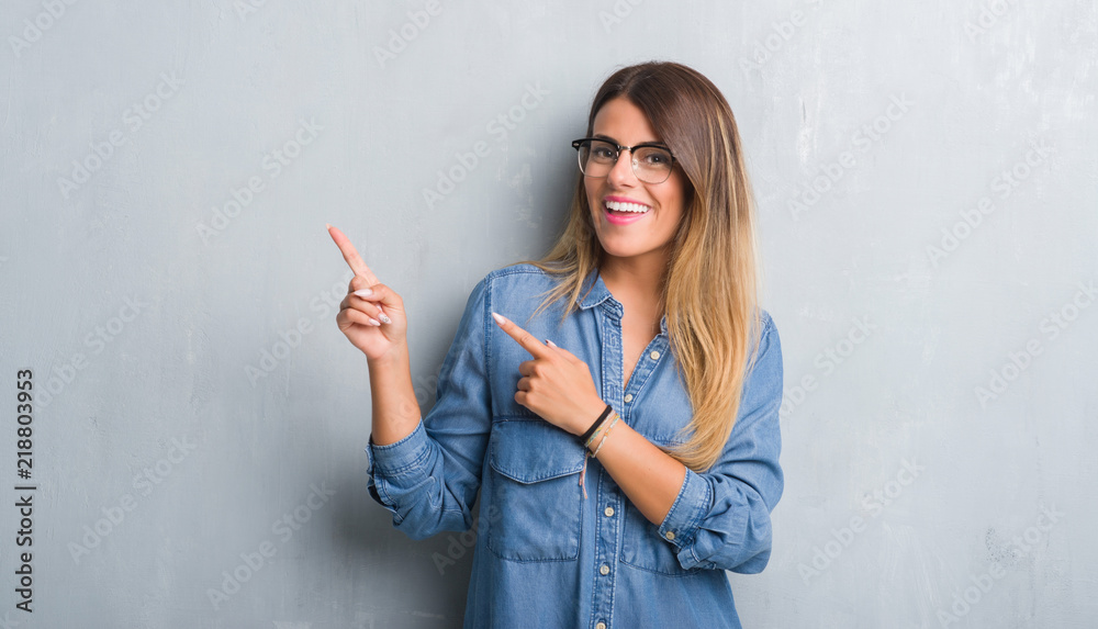 Young adult woman over grunge grey wall wearing glasses smiling and looking at the camera pointing with two hands and fingers to the side.
