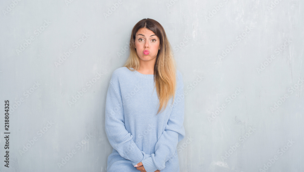 Young adult woman over grey grunge wall wearing winter outfit puffing cheeks with funny face. Mouth inflated with air, crazy expression. Young adult woman over grey grunge wall wearing winter outfit puffing cheeks with funny face. Mouth inflated with air, crazy expression.