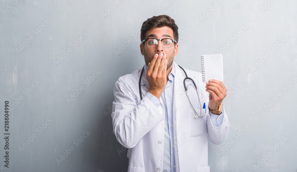 Handsome young doctor man over grey grunge wall holding blank notebook cover mouth with hand shocked with shame for mistake, expression of fear, scared in silence, secret concept Handsome young doctor man over grey grunge wall holding blank notebook cover mouth with hand shocked with shame for mistake, expression of fear, scared in silence, secret concept