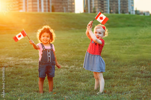 Happy adorable little blond Caucasian and hispanic latin girsl smiling holding hands and waving Canadian flags in park outdoors. Multiracial children celebrating Canada Day. Flag Day concept.