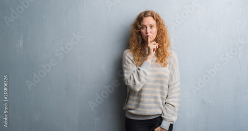Young redhead woman over grey grunge wall asking to be quiet with finger on lips. Silence and secret concept.
