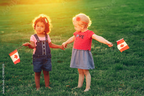 Happy adorable little blond Caucasian and hispanic latin girsl smiling holding hands and waving Canadian flags in park outdoors. Multiracial children celebrating Canada Day. Flag Day concept. photo