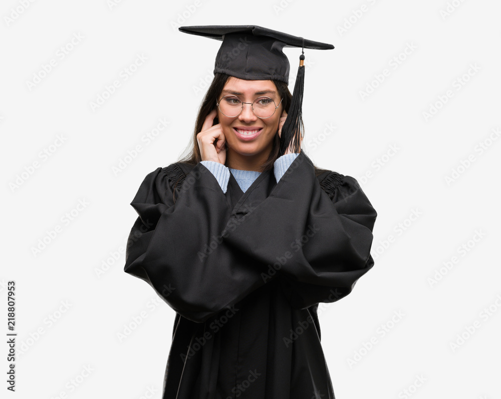 Young hispanic woman wearing graduated cap and uniform covering ears with fingers with annoyed expression for the noise of loud music. Deaf concept. Young hispanic woman wearing graduated cap and uniform covering ears with fingers with annoyed expression for the noise of loud music. Deaf concept.