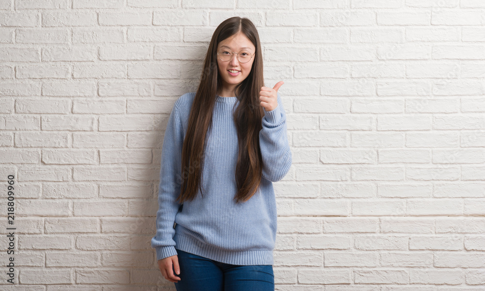Young Chinise woman over white brick wall doing happy thumbs up gesture with hand. Approving expression looking at the camera with showing success. Young Chinise woman over white brick wall doing happy thumbs up gesture with hand. Approving expression looking at the camera with showing success.