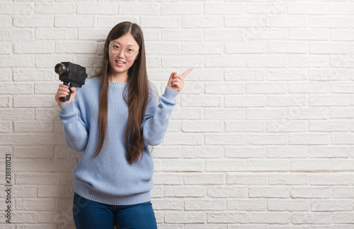 Young Chinese woman filming holding video camera very happy pointing with hand and finger to the side